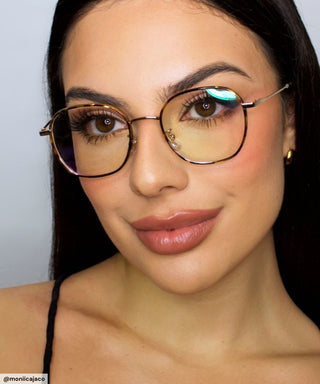 A woman with long dark hair, wearing vintage round EyeCandys Farrow Round glasses and a black top, smiles softly at the camera. She has subtle makeup, defined brows, and nude lipstick.
