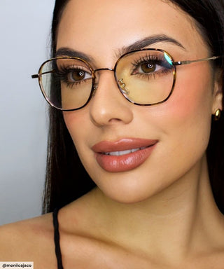 A woman with long dark hair and flawless makeup wears EyeCandys Farrow Round vintage gold-rimmed glasses. She pairs full lips, bold brows, and gold hoop earrings, gazing confidently at the camera.