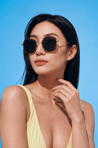 A woman wearing EyeCandys Farrow Round glasses and a light yellow top poses against a bright blue background, gazing slightly to the side with one hand on her chin.