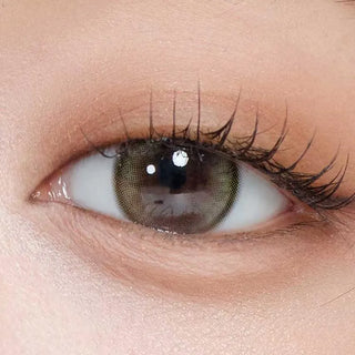 A close-up of a human eye with long lashes, smooth skin, and natural makeup wears Gemhour Demeter 1-Day 13.5 Olive Green contacts (10pk), showcasing the vibrant olive hue from Gemhour prescription color lenses.