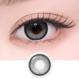 Close-up of an eye with long lashes wearing EyeCandys i-SHA Reborn Red 1-Day Milky Gray (10pk) colored contact lenses, with the lens design displayed below. Features a soft milky gray pattern with subtle depth for a natural, bright-eyed look in a comfortable silicone hydrogel daily lens.