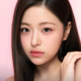 A young woman with long, straight dark hair and fair skin gazes at the camera with a neutral expression against a soft pink background. She wears subtle makeup and is seen wearing i-Sha One Pick Only One 1-Day Cherry Choco contact lenses by i-Sha, which give her eyes a natural dolly effect as she holds her hand near her face.