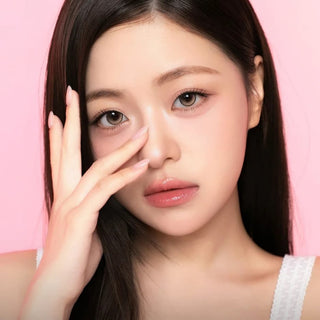 A young woman with long dark hair touches her face and gazes at the camera with a neutral expression. She has flawless skin, subtle makeup, and wears i-Sha One Pick Only One Dusty Brown contact lenses by i-Sha, which feature a 3-tone gradient design for natural enhancement. The soft pink background complements her look.