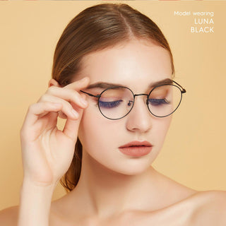 A young woman with light skin and straight brown hair wears EyeCandys Luna Round titanium glasses, posing with eyes closed against a beige background. "Model wearing LUNA ROUND" is displayed in the upper right corner.