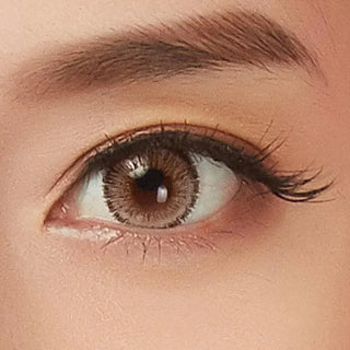 A close-up of an eye with a brown iris, enhanced by NEO Vision Glamour Brown (Custom Toric for Astigmatism) lenses. Long lashes, a defined brow, and smooth skin highlight the upward glance.