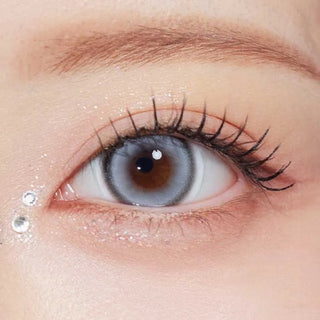 A close-up of an eye wearing Olola Glint 1-Day Grey (10pk) lenses by Olola, featuring long lashes, sparkly makeup, and two rhinestones with glitter at the outer corner for a dazzling look.