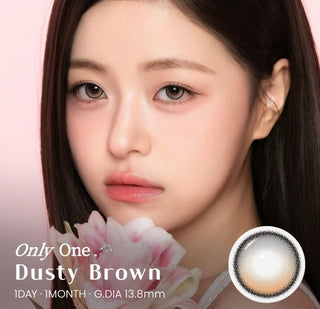 A woman with flawless makeup and straight brown hair holds a pink flower, beautifully highlighting her naturally enhanced eyes with i-Sha One Pick Only One 1-Day Dusty Brown (10pk) contact lenses by i-Sha. Text reads: “i-Sha One Pick Only One Dusty Brown 1-Day | G.DIA 13.8mm.” A close-up in the corner showcases the daily disposable colored contacts.