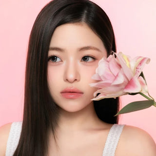A young woman with long, straight dark hair holds a pink lily flower near her face. She has clear skin, natural makeup, and wears a white top. Her i-Sha One Pick Only One Velvet Smoke Grey contacts by i-Sha add a subtle elegance to her look, perfectly complementing the soft pink gradient background.