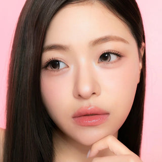 A young woman with long straight dark hair and flawless, glowing skin poses against a pink background, gazing off-camera with a neutral expression and her hand resting gently near her chin. She wears i-Sha One Pick Only One 1-Day Cherry Choco contact lenses by i-Sha, which give her eyes a natural dolly effect and enhance her look with subtle elegance.