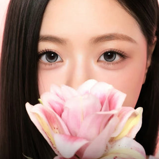 A woman with long, straight dark hair holds a large pink and white flower near her face, partly covering her mouth and nose. Her eyes, wearing i-Sha One Pick Only One 1-Day Velvet Smoke Grey (10pk) by i-Sha, gaze directly at the camera.