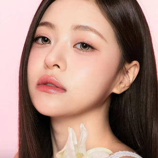 A young woman with long, straight dark hair and flawless skin gazes at the camera against a pink background. She wears soft makeup, glossy lips, and has a white flower near her shoulder. Her eyes are subtly brightened by i-Sha One Pick Only One Dusty Brown contact lenses from i-Sha, featuring a 3-tone gradient design for natural enhancement.