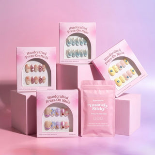 Four boxes of EyeCandys Main Stage Press-On Nail Sets and one pink packet of "Insanely Sticky" nail tabs are arranged on pastel pink blocks, with a soft pink backdrop, creating the look of a salon gel manicure right at home.