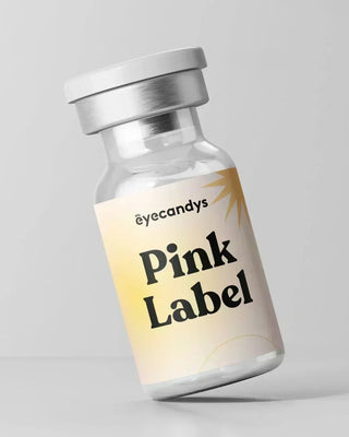 A small glass vial with a metallic cap bears the "EyeCandys" name and "Pink Label Vintage Grey" on a pink-yellow gradient label. The bottle, holding Vintage Grey color contact lenses for a dolly look, rests tilted on a plain grey backdrop.