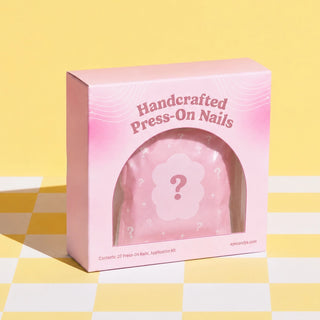 Handcrafted Press-O Nails mystery blind box in a pink cosmetic box with a frosted mystery pouch visible through an arched window on a pastel yellow checkered background.