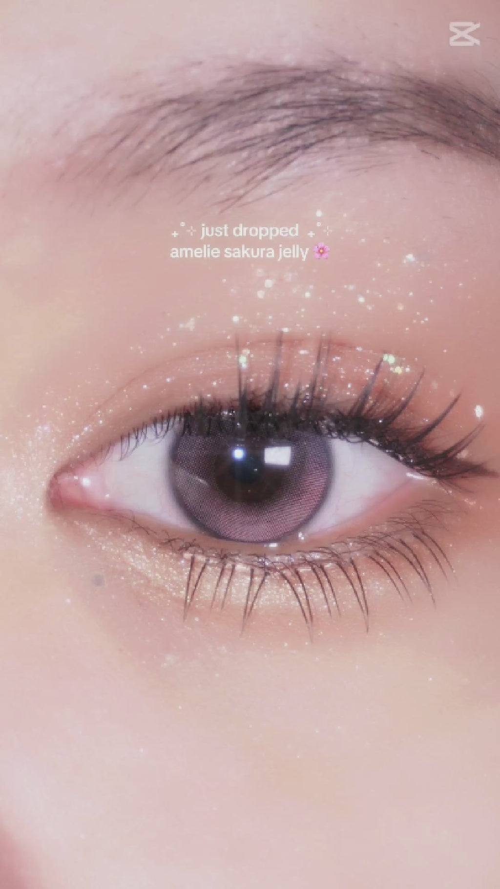 Model with long lashes and sparkly eye makeup applying EyeCandys Amelie Sakura Jelly colored contact lens using a contact lens applicator. The lenses feature a delicate sakura-inspired design, adding a soft pink tone for a vibrant, natural look. Perfect for those seeking colored contact lenses for dark eyes or a trendy, subtle enhancement. 
