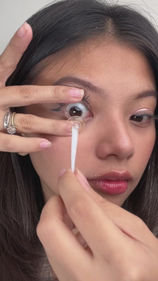Model applying EyeCandys Glossy Hazel colored prescription contact lens with a contact lens applicator, showcasing the difference between her eyes with and without the contact lens. The colored contact lens enhances her natural eye color with a warm, hazel tone, ideal for those seeking natural looking colored contacts. Perfect for individuals needing prescription contact lenses, these daily lenses provide comfort and a subtle, beautiful enhancement for dark eyes. Ideal for anyone looking for prescription co