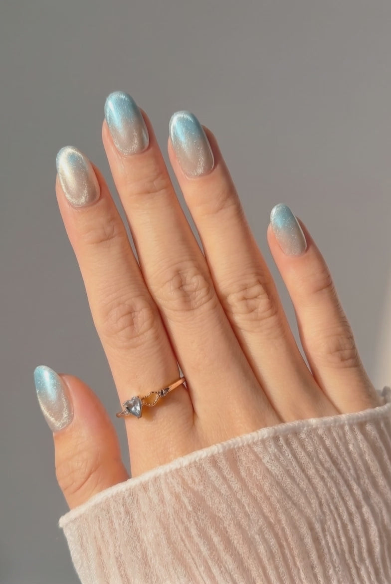 Wearing EyeCandys Moon Glass Press-On Nails, cool blue cat-eye gradient at the tips fading into a soft milky base with chrome glaze in an oval shape