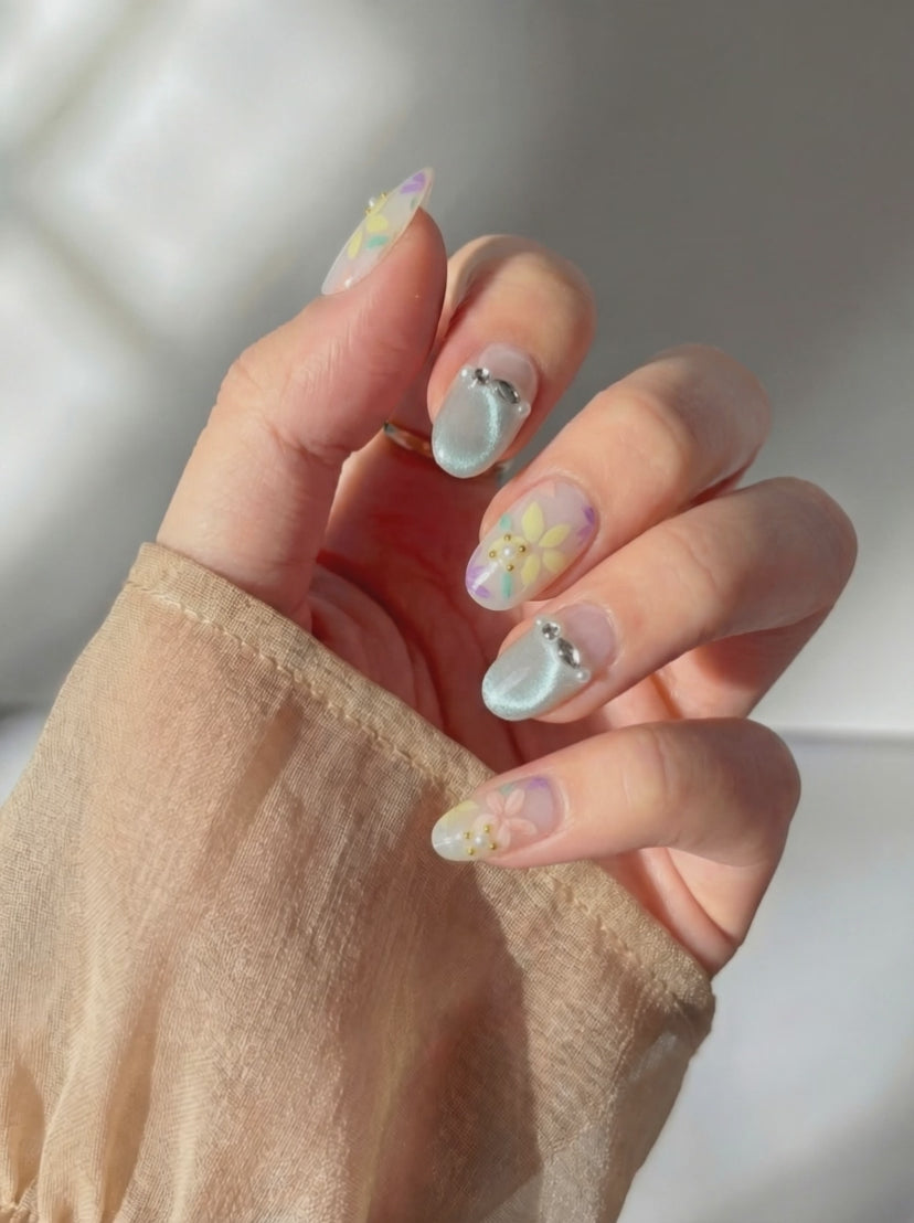 Wearing Eyecandys Floreline press on nails, featuring a sheer milky base with hand-painted floral designs in yellow, lavender, peach, and mint, plus soft shimmer accents in a short oval shape