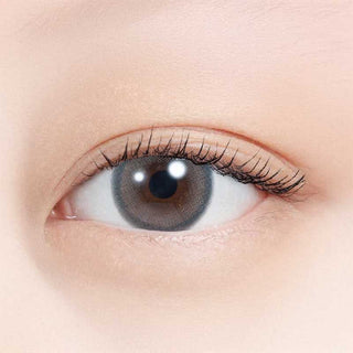 A close-up of a human eye wearing N's Collection Sakana Grey (10pk) colored contacts by N's Collection, with long eyelashes and natural makeup. The smooth, light-toned skin highlights the daily disposable contacts for myopia.