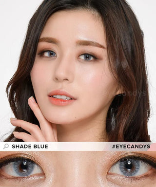 A woman with long dark hair and natural makeup wears EyeCandys Shade Blue contacts, highlighting her vivid blue eyes. She gently touches her face in a close-up. Text reads “SHADE BLUE” and “#EYECANDYS.”.